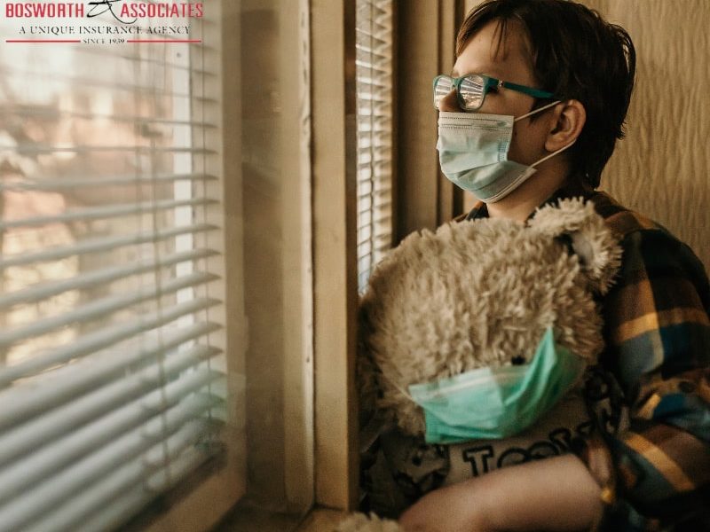 An old lady with glasses and a protective mask stands in front of the window in her home, holding a teddy bear with a protective mask during the Coronavirus lockdown 2020.