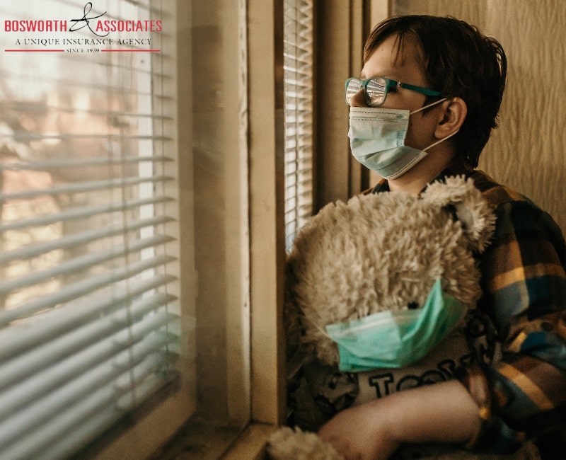 An old lady with glasses and a protective mask stands in front of the window in her home, holding a teddy bear with a protective mask during the Coronavirus lockdown 2020.