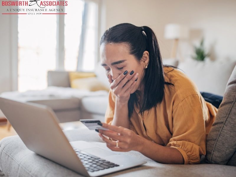 A woman in a yellow t-shirt holding her card in front of the laptop, looking upset. Possible victim of an insurance scam.