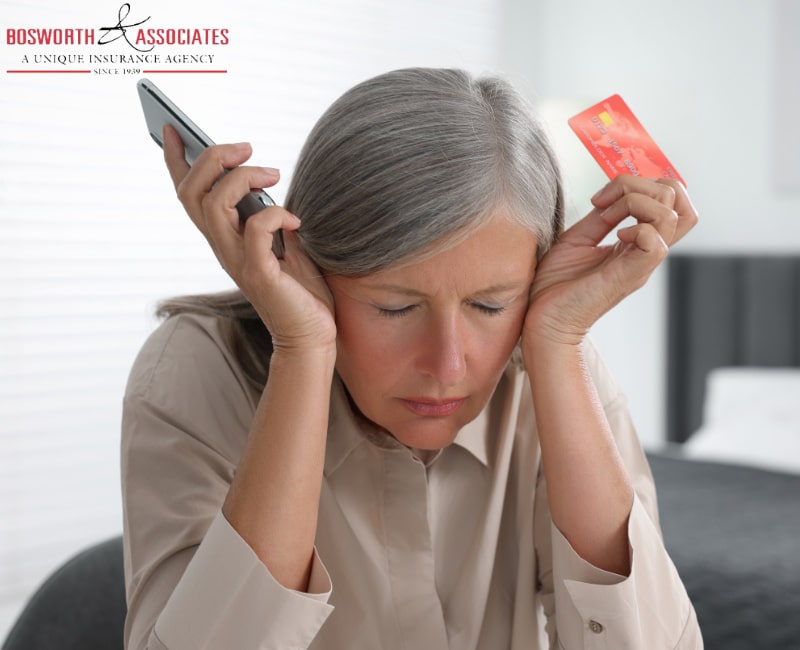 A woman in a gray car holds a credit card and smartphone. She has closed her eyes and looks upset after experiencing an insurance scam.